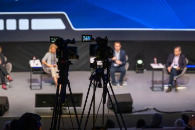 Sahnedeki panel tartışması. Kameralar seyirciyi çekiyor ve canlı olay aydınlanıyor.
