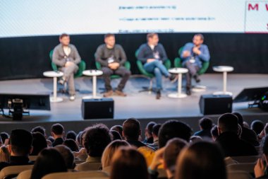 Canlı konferans sırasında sahnede dört konuşmacı paneli, yeşil sandalyeler, yuvarlak masalar, dikkatle dinleyen seyirciler, profesyonel etkinlik