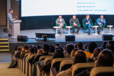 Seyirciler konferansta sahnede dört konuşmacı, mikrofonlar, büyük ekran ve ilgili katılımcılarla panel tartışmasını izliyor