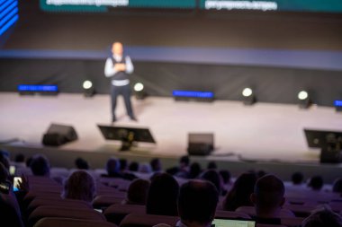 Bulanık konferans sahnesi sunucu, parlak ışıklar, büyük salonda izleyiciler, katılımcılar dinamik olay atmosferi yaratan cihazlarla etkileşim içindeler.