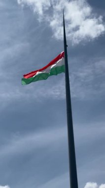 Macar bayrağı, açık mavi bir gökyüzünün altında uzun bir bayrak direğinin üzerinde dalgalanırken, yumuşak bulutlar, vatanseverlik, açık hava sembolizmi, ulusal kimlik ve kamuoyu önünde dinamik rüzgarı yakalıyor..