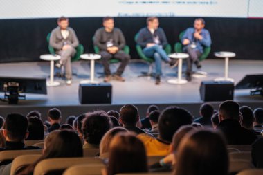 Konferans ortamında dört konuşmacı, dikkatle dinleyici, panel tartışması ve diyalog, profesyonel olay atmosferi