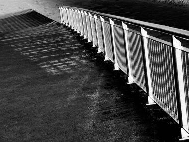 Siyah ve beyaz görüntü, güçlü çapraz hatları ve ıslak kaldırımda desenli gölgeleri olan ve çarpıcı şehir havası ve derinlik hissi yaratan yürüyüş yolunun yanında metal korkuluklar içerir..