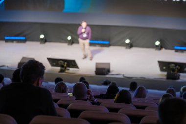 Konferans sırasında seyircilere hitap eden bulanık sahne konuşmacısı. Parlak ışıklandırma arka plan ekranı. Katılımcılar oturmuş performansı izliyorlar.
