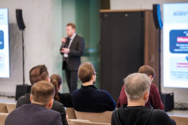 Sahnedeki konuşmacı ile profesyonel konferans sahnesi seyirciyi ileriye dönük olarak bulanıklaştırarak sunumu dinliyor. Geniş ekran slaytları modern iş atmosferi yaratıyor