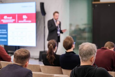 Sunucu, şirket konferansı sırasında oturma izleyicilerine hitap eder. Geniş ekran grafikleri gösteriyor. Camdan duvarları olan modern bir oda profesyonel atmosfer yaratır. Katılımcılar, dinleyin ve not alın..