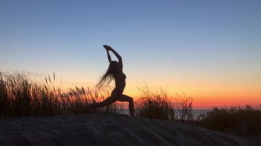 Gün batımında kumlu sahilde uzun otlar ve sakin ufuklarla yoga yapan birinin silueti. Sakinlik sahnesi denge esnekliği ve gevşemeye odaklı..