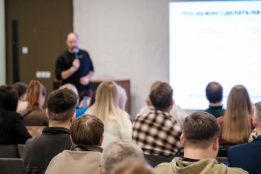Canlı konferans salonu sahnesi, seminer sırasında canlı ekranla izleyicilere hitap eden sunucuyu gösteriyor. Profesyonel atmosfer öğrenme, işbirliği ve iş iletişimini vurguluyor.