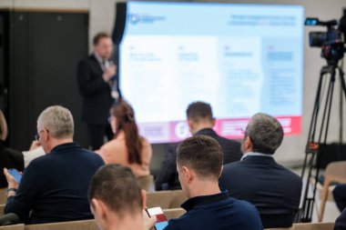 Seyirciler konferansa büyük ekranda sunum yapan bir konuşmacı olarak katılıyor. Kamera ekibi olayı modern bir mekanda kaydediyor..