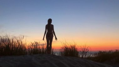 Gün batımında kumlu kumulda bikinili bir kadın silueti. Sıcak, huzurlu plaj sahnesi. Rahatlama özgürlüğü için ilham verici..