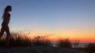 Gün batımında sahilde yürüyen genç bir kadının silueti. Sıcak turuncu gökyüzü, sakin deniz çimenleri üzerinde sallanıyor ve sakin bir sahil havası yaratıyor..