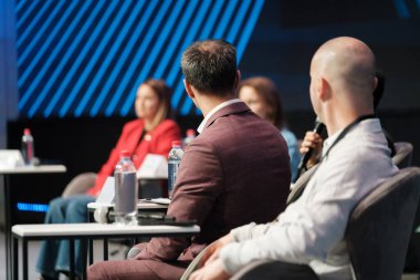Panel discussion at conference with professionals listening and speaking on stage. Attendees wear business attire, water bottles on tables, microphones in use.