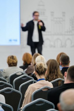 İş konferansında sunucuyu dinleyen seyirciler, ön planda iple odaklanmış katılımcı, sahnede bulanık hoparlör, profesyonel etkinlik atmosferi.