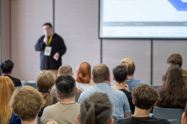 Konferans izleyicileri, sunum sırasında ekranlı parlak odada konuşmacı dinlerler.