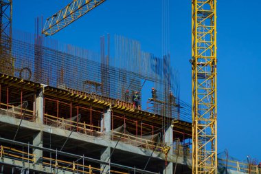 İnşaat işçileri yüksek katlı beton binaya çelik inşaat demirleri döşüyorlar, açık mavi gökyüzüne karşı sarı vinç..