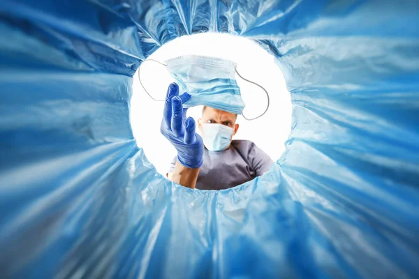 A blue-gloved man throws a used medical mask in the trash, a view from the bucket. Concept of medical waste disposal and consequences of virus spread protection