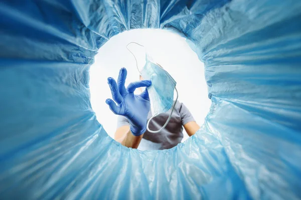 A blue-gloved man throws a used medical mask in the trash, a view from the bucket. Concept of medical waste disposal and consequences of virus spread protection