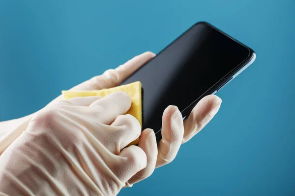 Hands in protective gloves clean and disinfect a smartphone with a yellow napkin on a blue background. Prevention of viral infections. Close-up