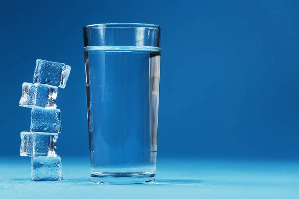 Ice cubes tower with a glass of cold and clean water on a blue background. Free space