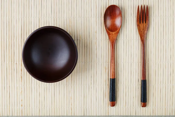Empty dark wooden cup, spoon and fork made of natural wood on a light bamboo backing. View from above