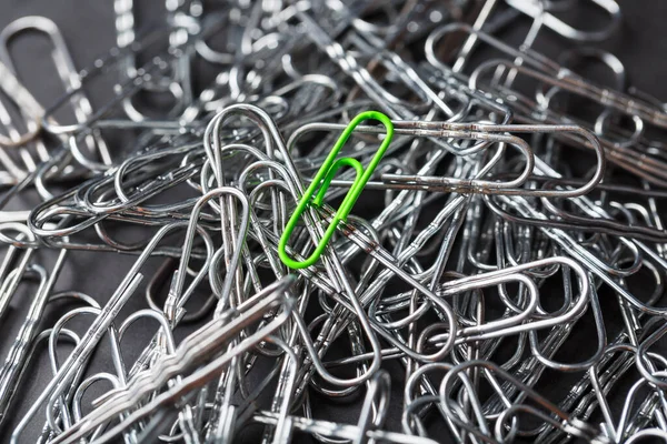 The green paper clip stands out against a textured background of silver paper clips. Concept