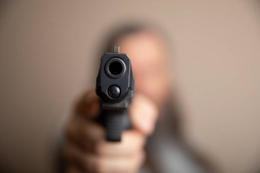 Bir kişi bir silah tutuyor ve kameraya doğrultuyor. Tehlike ve korku kavramı, kişinin eline silah alıp doğrudan izleyiciye bakması gibi. Manzara gergin ve rahatsız edici.
