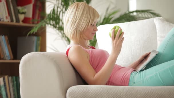 Meisje op sofa met touchpad eten apple — Stockvideo