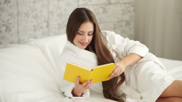 Girl in bathrobe lying in bed reading book — Stock Video