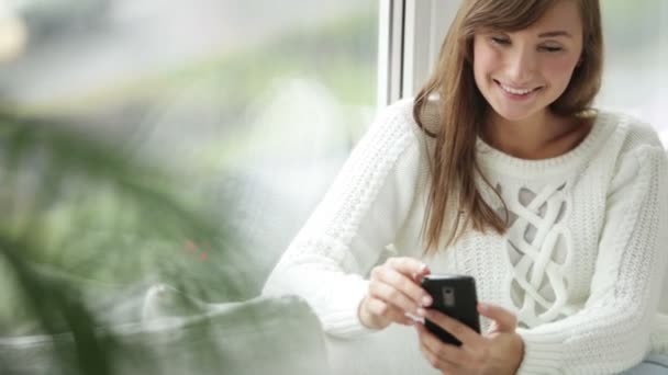 Девушка сидит у окна с помощью мобильного телефона — стоковое видео