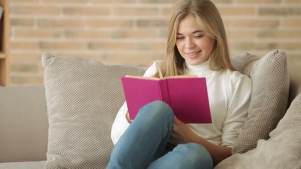 Девушка сидит на диване и читает книгу — стоковое видео