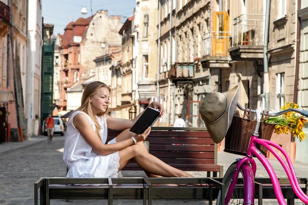 Girl with bicycle sitting on bench and reading book — Stock Photo, Image