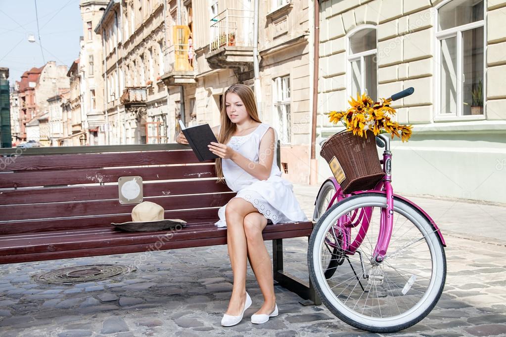 girl with bicycle sitting on bench and reading book