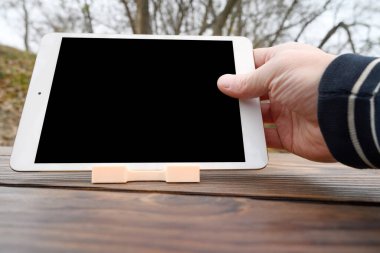 Bir el beyaz bir tablet tutuyor, yatay olarak temiz siyah bir ekranı var, kahverengi ahşap bir masanın üzerinde küçük bir ışık tutacağı var.
