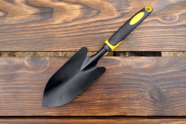 Bitkilerin nakli ve bakımı için tasarlanmış parlak sarı ve siyah bir kulpu olan siyah bahçe malası, desenli kahverengi ahşap bir yüzeye dikkatlice uzanarak sonbahar baharı bahçe aletlerini gösteriyor.