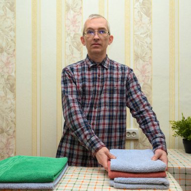 Gözlüklü ve kareli gömlekli bir adam masanın arkasında duruyor ve düzgünce katlanmış bir havlu yığını yerleştiriyor, çamaşır ve ütü işlemini tamamlıyor ve evdeki düzeni koruyor.