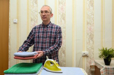 Gözlüklü ve kareli gömlekli bir adam ütüledikten sonra rahat bir odada duruyor, düzgünce katlanmış bir havlu yığını tutuyor ve onları dolaba koymaya hazırlanıyor.