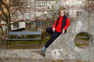 Kırmızı yelekli ve koyu renk elbiseli bir çocuk bir apartmanın avlusunda eski bir beton blokta oturuyor. Sonbahar günü, ön manzara, arkada bir bank ve ağaçlar.