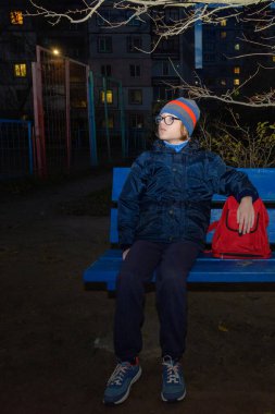 Gözlüklü yalnız çocuk günün karanlık saatlerinde oyun alanında dinleniyor, arka planda çok katlı binanın pencereleri parlıyor, çocuk ailesini bekliyor ve ilgiyle bakıyor.