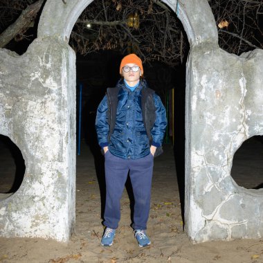 Turuncu şapkalı çocuk beton deliğin içinde poz veriyor, ellerini cebinde tutuyor ve kendinden emin bir şekilde ileri bakıyor, etrafında yaşlı park ağaçlarının karanlık ve dalları var.