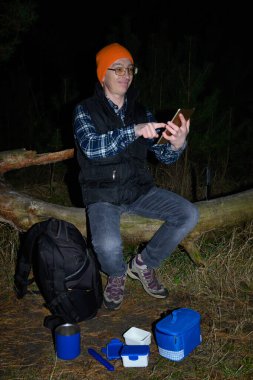 Sırt çantasının ve yemeğin yanında dinlenen gülümseyen gezgin geceleri karanlık orman ortamında gezinme veya eğlence için tablet bilgisayar kullanırken