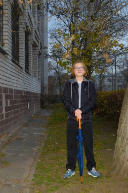 Ciddi bir ifadeye sahip genç, bir sonbahar ağacının altında çimenlikte duruyor. Sarı yapraklar, alacakaranlıkta çok katlı bir binanın arka planına karşı kamış şemsiyesine yaslanıyor.