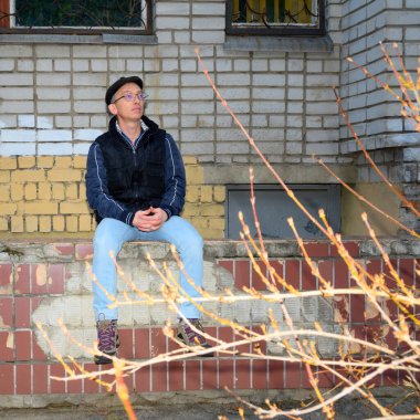 Gözlüklü ve kalın elbiseli yetişkinler sokakta alçak bir duvarda otururken güneşli bir ilkbaharda ön planda bulanık dallar görülebilir.