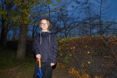 Gözlüklü bir gencin portresi ve çimlerin üzerinde sarı yapraklı bir ağaç gövdesinin yanında duran koyu renk ceketli katlanmış mavi bir şemsiyenin parlak turuncu sapını tutan