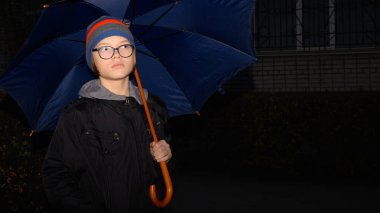 Gözlüklü bir genç, çizgili bir şapka ve siyah bir ceket, alacakaranlıkta tuğla duvara bakan ahşap saplı mavi bir kamış şemsiyesinin altında duruyor.