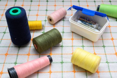 Dikiş makinesi ve el dikimi için çok renkli iplikler, profesyonel terzilik için keskin iğneler ve iğneler içeren kutunun yanındaki açık masa örtüsüne saçılmıştır.