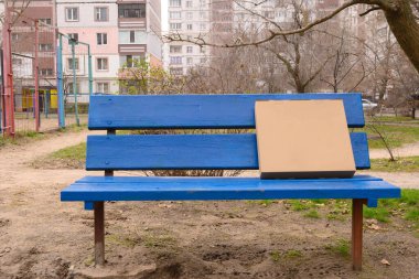 Açık kahverengi bir karton kutu, yüksek katlı bir binanın avlusunda, parlak mavi bir ahşap bankta duruyor.