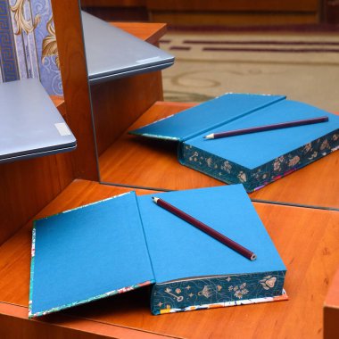 Üzerinde mavi sayfalar ve koyu kırmızı bir kalem bulunan açık bir defter. Aynanın önünde, tahta bir yüzey üzerinde duruyor. Kapalı gri bir dizüstü bilgisayarın kenarı odada solda görünür durumda.