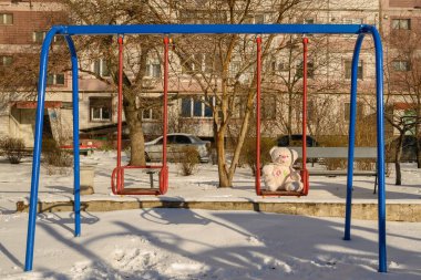 Mavi metal salıncaklı, kırmızı koltuklu beyaz bir oyuncak ayı karlı bir oyun parkında oturuyor kışın çok katlı bir eve ve çıplak ağaçlara karşı.