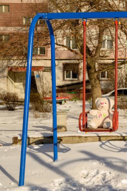 Beyaz ayıcık kırmızı bir salıncak koltuğunda oturuyor, beyaz kar etrafta uzanıyor, ağaçlar ve bir bina cephesi güneşli bir kış gününde arka planda görünür.