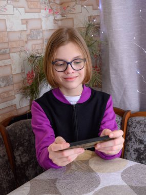 Gözlüklü ve mor ceketli genç adam mutfak masasında oturur ve telefon ekranına gülümser. Ev ortamında iletişimin ya da mobil bir oyunun tadını çıkarır.